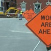 Close up of construction signs on side of road, with flagger and heavy equipment in background.