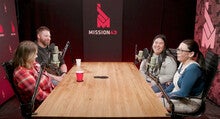 Four people sit at a table in front of microphones and talk while recording a podcast.