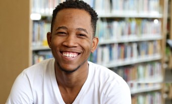 Happy student from Jamaica wearing a white t-shirt sitting in front of shelf of books