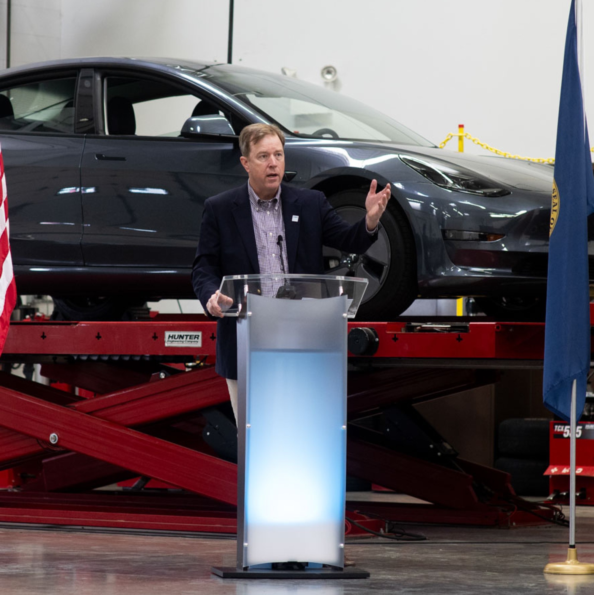 The College president speaks from a podium in front of a newly delivered Tesla electric vehicle.