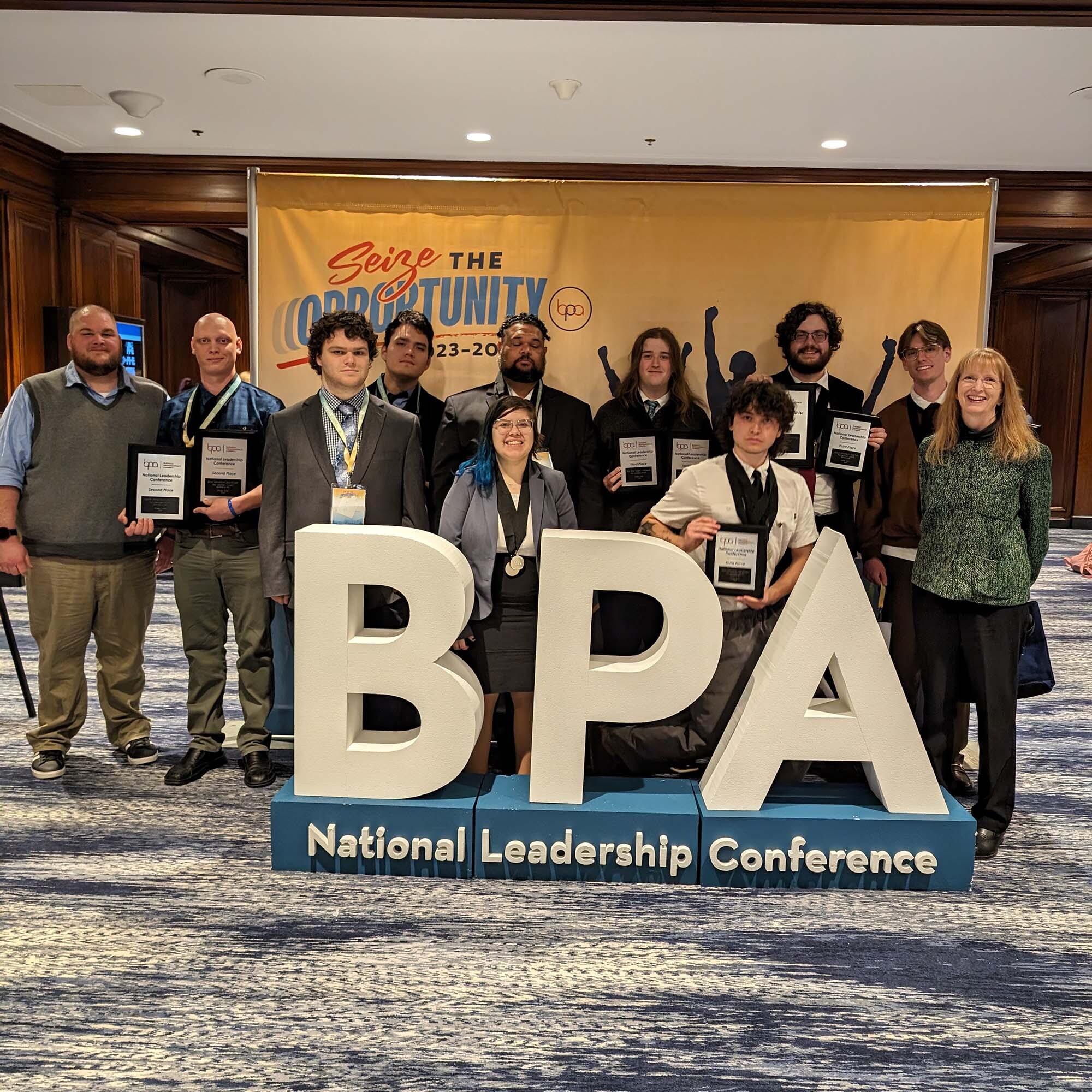 Student from CWI smile and hold award plaques behind large letters that say "BPA, National Leadership Conference."