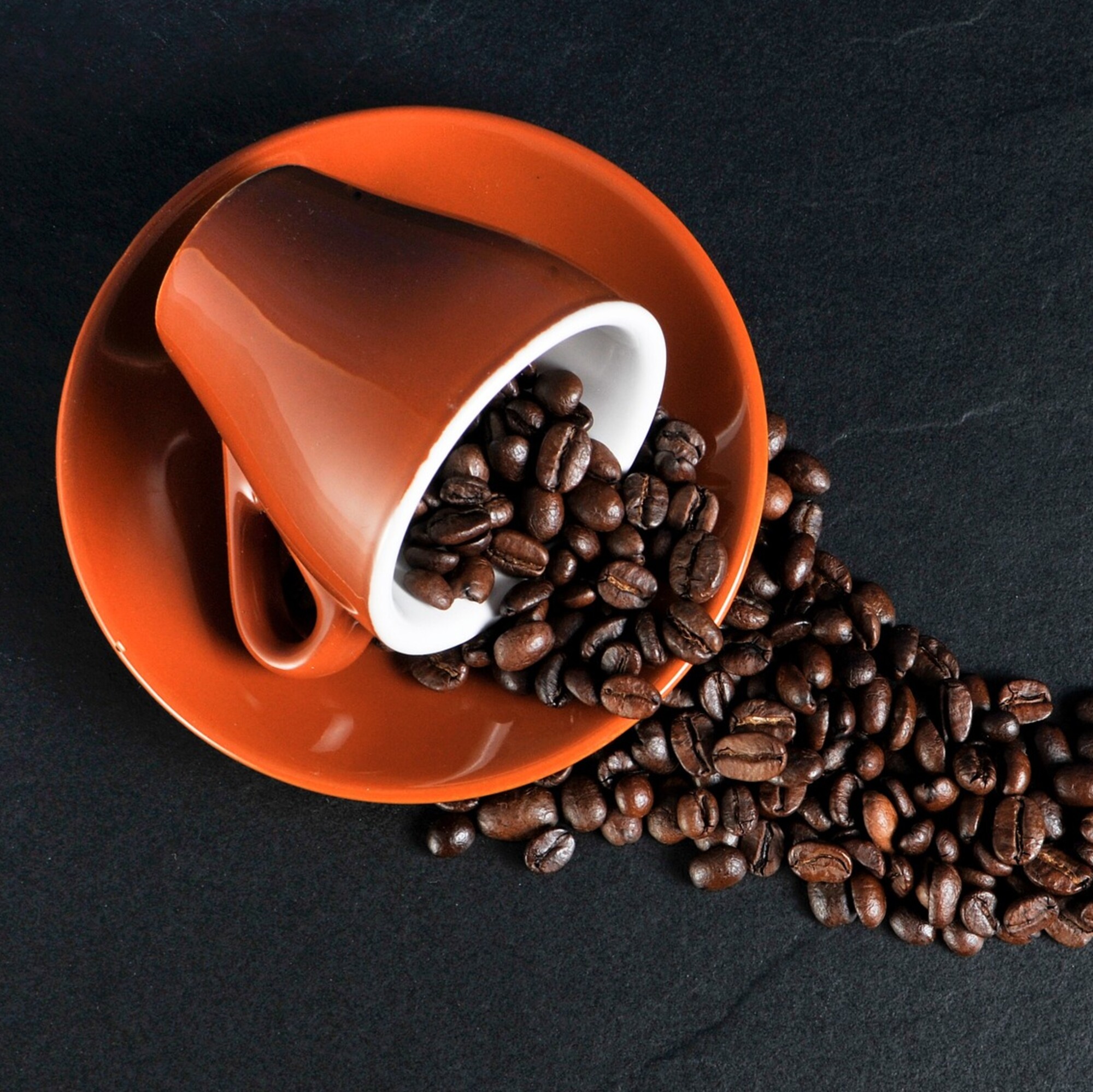 A red coffee cup rests on it's side on a black table with coffee beans spilling out.