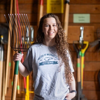 A student stands in a barn holding a pitchfork. 