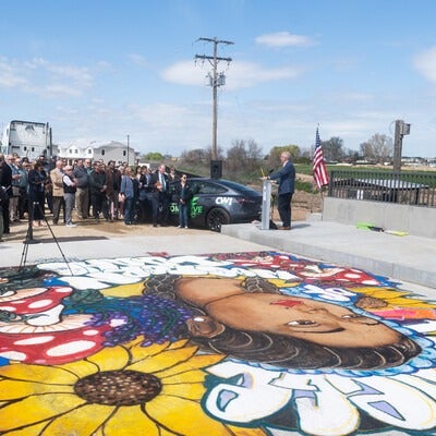 A large group of people listen to a man addressing the crowd, standing in front of a newly constructed bridge.