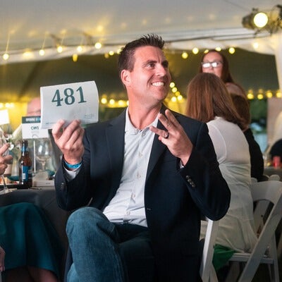 Person raising a bid paddle with the number 481 during an auction event, with string lights and attendees in the background.