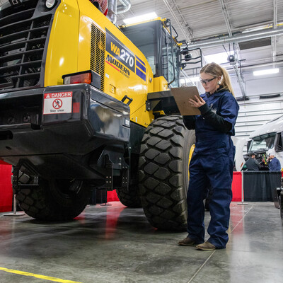 A student wearing coveralls holds a clipboard while inspecting a yellow tractor.