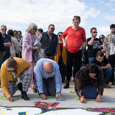 Several people kneel down on concrete to sign their names in paint pen on a newly constructed bridge.