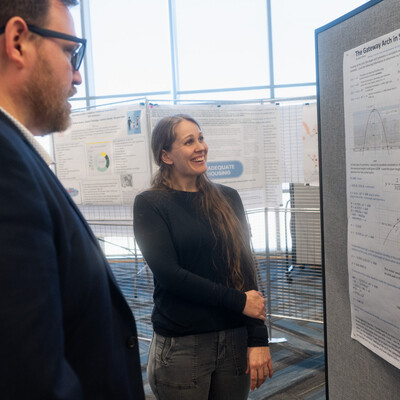 A student smiles as she discusses research, displayed on a hanged poster, with a faculty member.