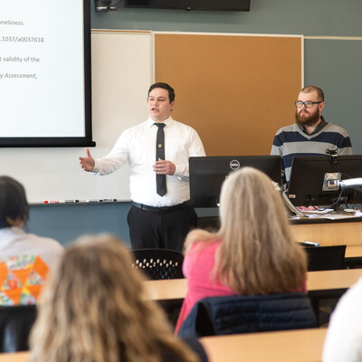 Two students stand at the front of a classroom presenting a mini-talk about a research project, which is displayed on a screen.