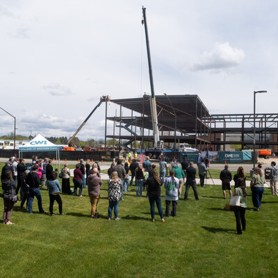 An audience listens to a speaker outdoors in front of a construction site on a college campus.