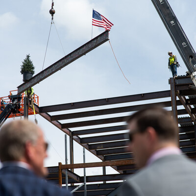 Two men watch from the ground in front of a construction site as a metal beam is lowered onto a buildings frame.
