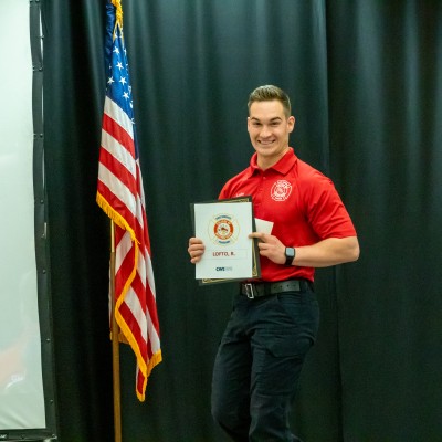 A graduate smiling with a certificate in front of an American flag.