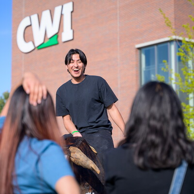 A student laughs while riding a mechanical bull while watched by friends. The CWI logo is on a building in the background.