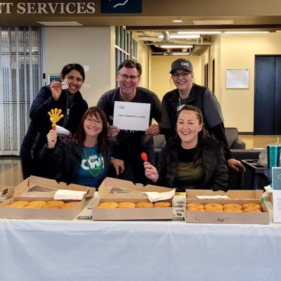 3 smiling staff members hold a white sign that says "CWI changes lives" in front of a table of donuts.