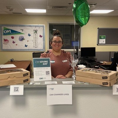 A smiling staff member smiles at the camera in front of signs that say "CWI Giving Day" and boxes of donuts.