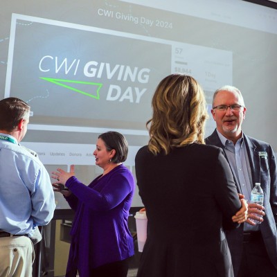 People mingle in a large room in front of a projected image on a screen that says "CWI Giving Day."