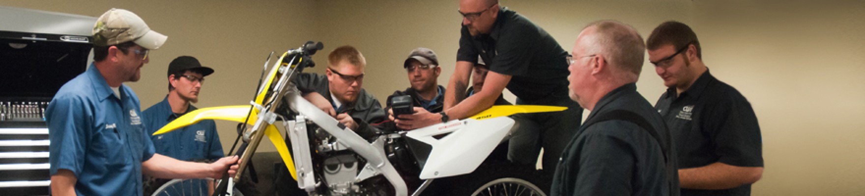 Students and instructors around a yellow and white motorcycle in a classroom.