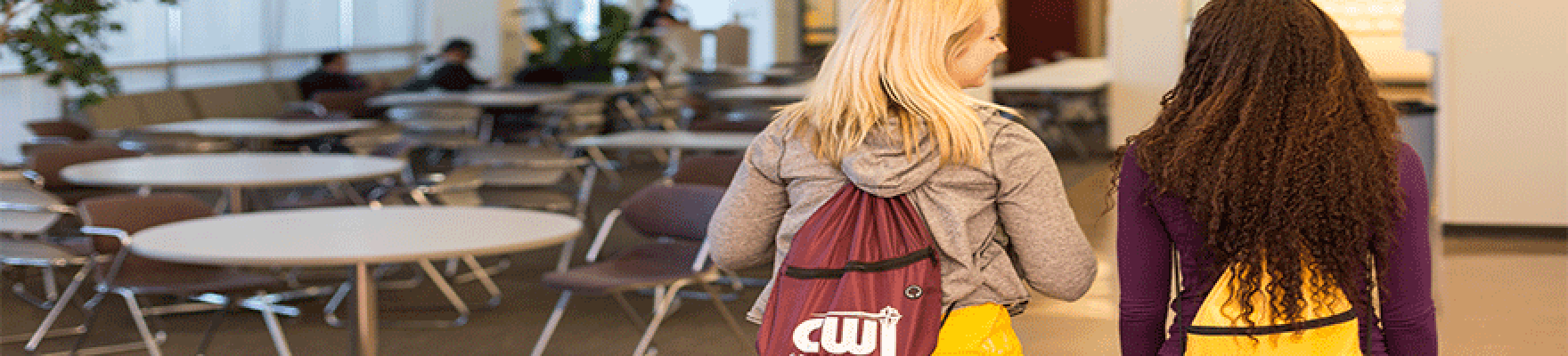 Two female students with CWI backpacks walking through common area with tables in background.