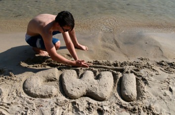 Male student spelling C W I in sand near water