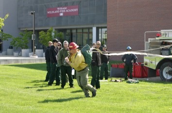 Wildland fire students pulling hoses across grass in front of a brick building.