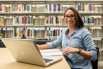 Student sitting in the library with a laptop and notebook smiling at the camera