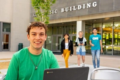 Student sitting outside a campus building with a laptop, students walking out of the building in the background