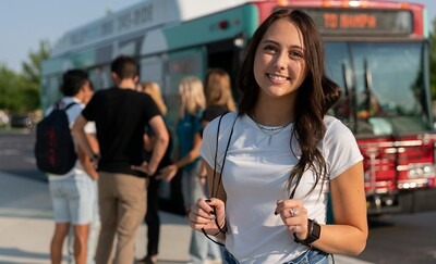 student standing near a bus with other students in the background wearing a backpack
