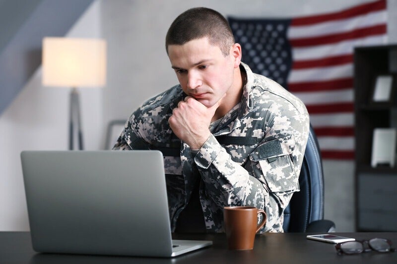Military student studying on a laptop at a desk with an American flag in the background