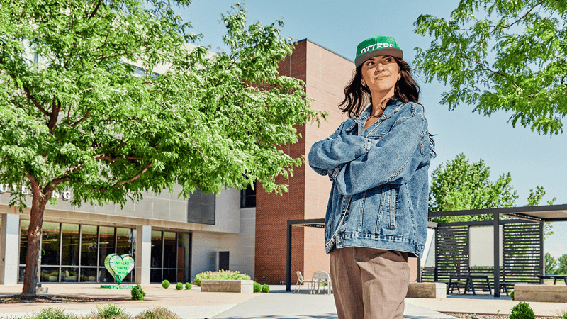 Student wearing a hat standing with their arms crossed outside a campus building. 