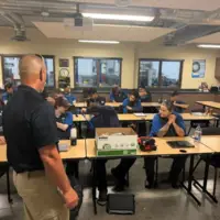 Instructor addresses students seated at tables in a classroom