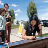 A girl signs her name onto a metal construction beam as two other stand watching her.