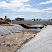 People stand on a newly constructed bridge, visible in profile from a distance as it crosses over a dry canal.