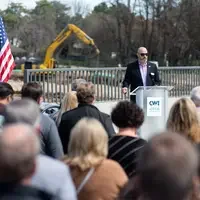 A man in sunglasses standing behind a lectern addresses a crowd of people at an out door event. 