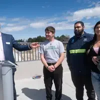 A speaker addressing an audience points to 3 people standing on his left.
