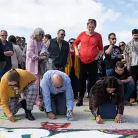 Several people kneel down on concrete to sign their names in paint pen on a newly constructed bridge.