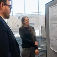 A student smiles as she discusses research, displayed on a hanged poster, with a faculty member.