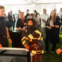 A young girl wearing firefighter equipment tests out a fire hose to put out a simulated fire.