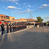 Group of firefighters and cadets standing in formation outside Nampa Fire Station during a ceremony, with fire trucks parked in 
