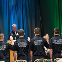 Students standing in front of chairs with their right hand raised 