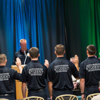 Students standing in front of chairs with their right hand raised 