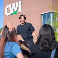 A student laughs while riding a mechanical bull while watched by friends. The CWI logo is on a building in the background.