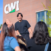 A student laughs while riding a mechanical bull while watched by friends. The CWI logo is on a building in the background.