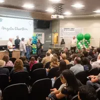 A graduating apprentice receives her certificate from instructors in a large classroom while an audience looks on.