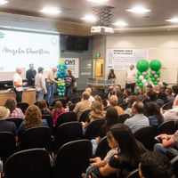 A graduating apprentice receives her certificate from instructors in a large classroom while an audience looks on.
