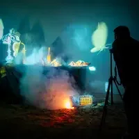A person stands by a tripod photographing a smoky, illuminated still life scene with assorted objects on a draped table and glow