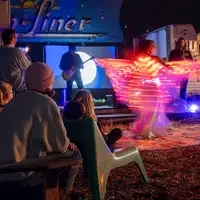 A group of people watch a nighttime outdoor performance with a musician on stage and a dancer twirling with bright LED lights.