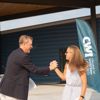Two people clasping hands in front of a "CWI College of Western Idaho" flag outside a building.