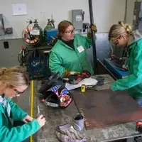 Three people in green jackets work on a metalworking project in a workshop with various tools and equipment around them.