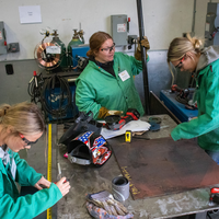 Three people in green jackets work on a metalworking project in a workshop with various tools and equipment around them.