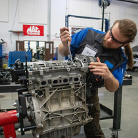 A person works on an engine in an automotive workshop with various tools and equipment around them.